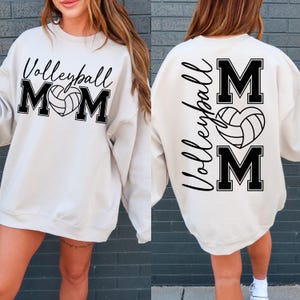 May include: A white sweatshirt with black text that says "Volleyball Mom" on the front and "Volleyball Mom" with a volleyball in a heart on the back.