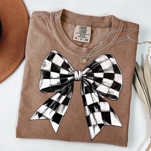 May include: A brown t-shirt featuring a black and white checked bow design. The shirt has a distressed look and the text "COMFORT COLORS" is visible on the tag.