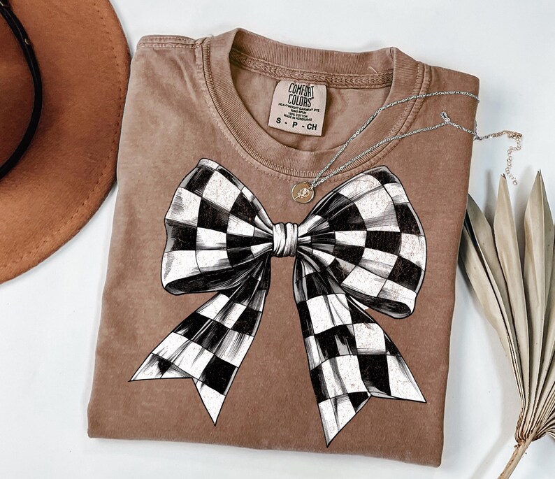 May include: A brown t-shirt featuring a black and white checked bow design. The shirt has a distressed look and the text "COMFORT COLORS" is visible on the tag.
