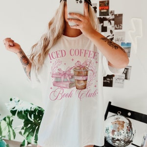 K&ouml;nnte beinhalten: Wei&szlig;es T-Shirt mit einer pinkfarbenen Grafik eines Stapels B&uuml;cher und einer Tasse Eiskaffee. Der Text "Iced Coffee Book Club" ist in pinker Kursivschrift geschrieben.