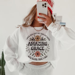 May include: White sweatshirt with a floral design and the text "I once was lost but now am found Amazing Grace Was blind, now I see".