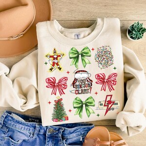 May include: Cream-colored sweatshirt with Christmas-themed graphics. The design includes a Santa Claus figure, green bows, red bows, a Christmas tree, string lights, and the text "YOU SERIOUS CLARE!".