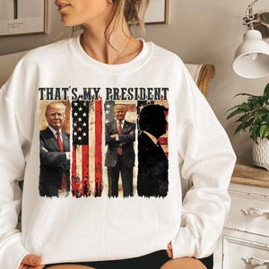 May include: White sweatshirt with a graphic print of three men in front of an American flag. The text "THAT'S MY PRESIDENT" is printed above the graphic.