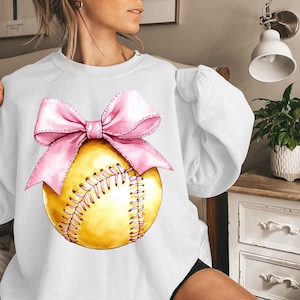 May include: White sweatshirt with a pink bow and a yellow softball with white stitching.