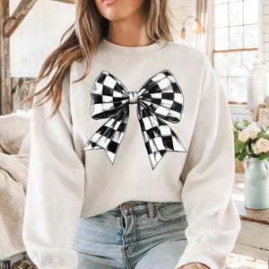 May include: A white sweatshirt featuring a black and white checked bow design.