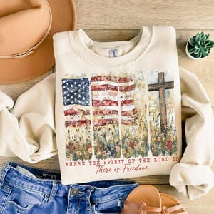 May include: A cream-colored sweatshirt with a watercolor design of an American flag and a cross. The text "Where the Spirit of the Lord is There is Freedom" is printed on the sweatshirt.