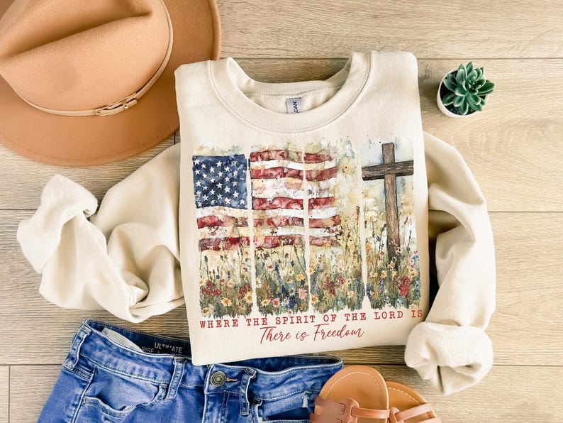 May include: A cream-colored sweatshirt with a watercolor design of an American flag and a cross. The text "Where the Spirit of the Lord is There is Freedom" is printed on the sweatshirt.