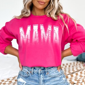 Op de afbeelding: Een roze sweatshirt met het woord "MAMA" gedrukt in witte, verweerde letters.