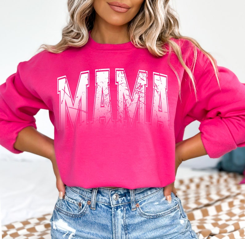 Op de afbeelding: Een roze sweatshirt met het woord "MAMA" gedrukt in witte, verweerde letters.