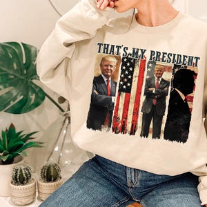 May include: A beige sweatshirt with a graphic print of three men in front of an American flag. The text "THAT'S MY PRESIDENT" is printed above the graphic.