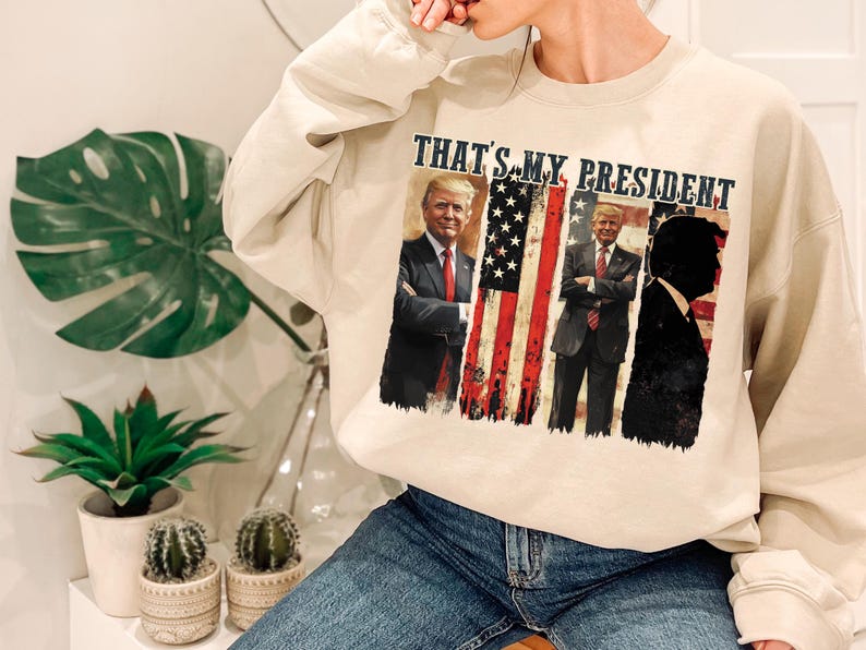 May include: A beige sweatshirt with a graphic print of three men in front of an American flag. The text "THAT'S MY PRESIDENT" is printed above the graphic.