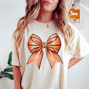 Pu&ograve; includere: T-shirt color crema con una grande grafica a fiocco arancione con contorni neri. Il disegno del fiocco &egrave; centrato sulla parte anteriore della maglietta. La maglietta ha maniche corte e una vestibilit&agrave; ampia.