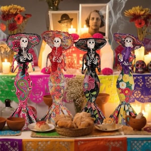 May include: Four standing skeleton figurines dressed in colorful patterned dresses and wide-brimmed hats. Each figure has a white skull face, skeletal hands crossed over the chest, and wears a vibrant outfit featuring floral, paisley, or botanical prints.
