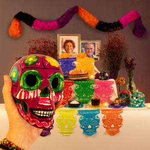 May include: A hand-held ceramic skull painted in vibrant magenta with green eye sockets, white teeth, and floral accents. The background features a Dia de los Muertos altar with colorful paper cutouts, framed photographs, and small decorative skulls.