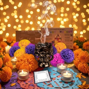 May include: A black ceramic incense burner emitting smoke, surrounded by orange and purple paper flowers, two lit tea lights in glass holders, and a small square card with the text Honoring My Family, set against a background of golden bokeh lights.