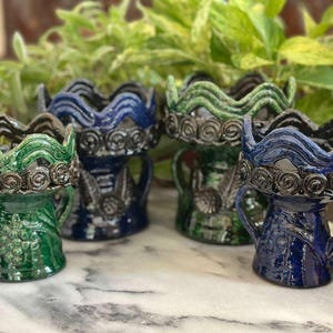 May include: Four ceramic candle holders in green and blue hues. Each has a wavy rim with a spiral design and two small handles. The holders are displayed on a marble-effect surface with greenery in the background.