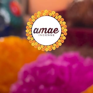 May include: A circular logo featuring the text "amae INCENSE" in dark brown lettering, centered within a wreath of vibrant yellow and orange marigold flowers. The background is softly blurred, showing out-of-focus orange and magenta floral elements.