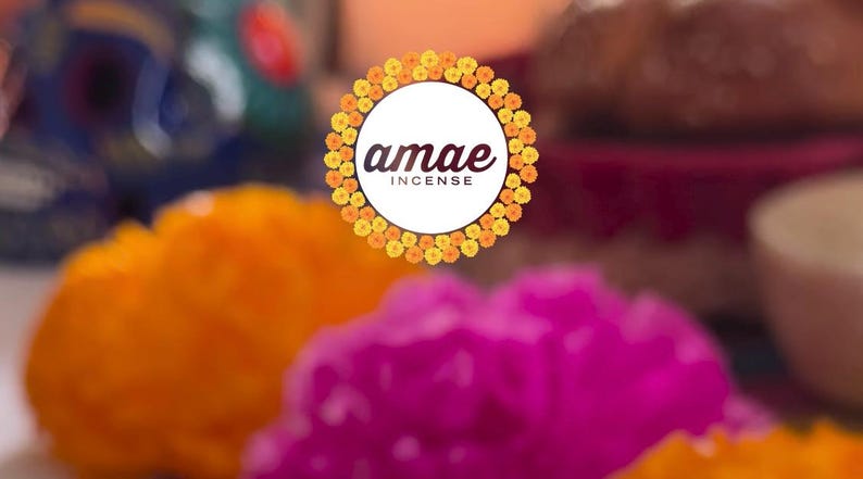 May include: A circular logo featuring the text "amae INCENSE" in dark brown lettering, centered within a wreath of vibrant yellow and orange marigold flowers. The background is softly blurred, showing out-of-focus orange and magenta floral elements.