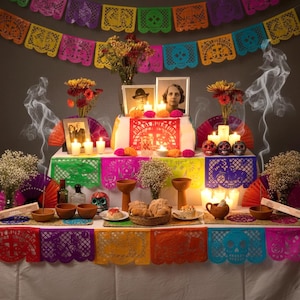 May include: A Dia de los Muertos altar featuring colorful papel picado banners, framed photographs, lit candles, marigolds, and baby's breath. The display includes ceramic skulls, clay bowls, bread, and smoking copal incense sticks on a white tablecloth.