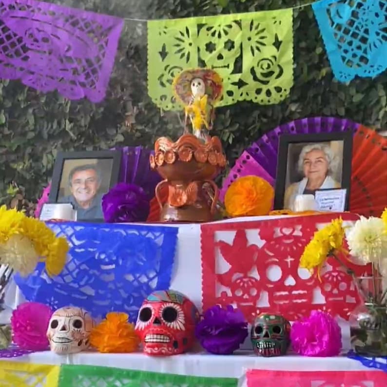 May include: A Day of the Dead altar featuring colorful papel picado banners, two framed portraits, a central clay incense burner, and three decorative skulls in white, red, and dark green, accented with bright yellow and purple paper flowers.