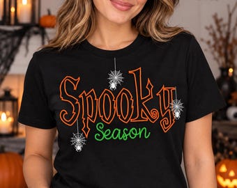 May include: A black short-sleeved t-shirt featuring the orange text Spooky and the green text Season. The design includes three white spider web graphics with small spiders hanging from the letters, creating a festive Halloween-themed graphic.