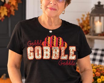 May include: A black short-sleeved t-shirt featuring the text Gobble Gobble in a decorative font with a patterned fill. Above the text is a stylized turkey tail graphic with red, orange, and yellow stripes. The design is embroidered or appliqued.