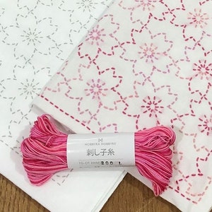 Può includere: Due quadrati di tessuto bianco con motivi floreali ricamati in filo grigio e rosa. Una bobina di filo variegato rosa e rosso con testo giapponese è davanti al tessuto. Il tessuto è probabilmente per progetti di ricamo o quilting.