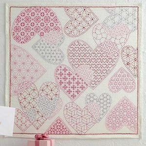 May include: A white fabric square with multiple pink and gray hearts stitched with different patterns. The hearts are arranged in a random pattern and overlap each other.