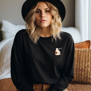 Könnte beinhalten: Ein schwarzes Langarm-Sweatshirt mit Rundhalsausschnitt und einer kleinen Grafik auf der linken Brust, die einen weißen Geist mit einem orangefarbenen Kürbis zeigt, sowie dem weißen Schriftzug BOO! auf dem schwarzen Stoff.