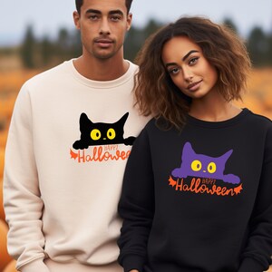 Könnte beinhalten: Ein beigefarbenes Sweatshirt mit Rundhalsausschnitt und einer schwarzen Katzengrafik mit gelben Augen sowie dem orangefarbenen Schriftzug Happy Halloween, und ein schwarzes Sweatshirt mit violetter Katzengrafik und gelben Augen.