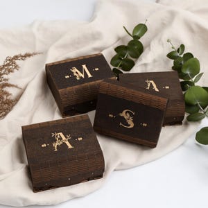 May include: Four dark brown wooden boxes with gold lettering and decorative accents. The boxes are arranged on a cream-colored cloth, with sprigs of greenery and dried plants. The letters "M", "A", and "S" are visible on the boxes.