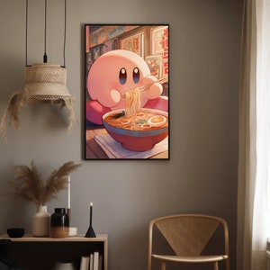 May include: A framed art print featuring a pink cartoon character eating ramen noodles with chopsticks. The character is seated in front of a red bowl filled with noodles, broth, and an egg. The background shows a wall with framed pictures.