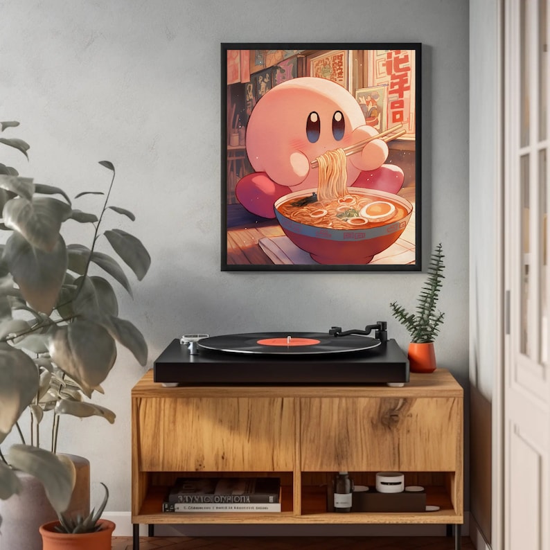 May include: A framed art print featuring a pink cartoon character eating ramen noodles with chopsticks. The character is in front of a bowl of ramen with an egg. The background shows Japanese text and a bookshelf. The print is hanging above a wooden cabinet.