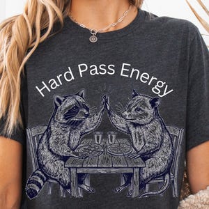 May include: Dark gray t-shirt featuring a graphic of two raccoons giving each other a high five over a table with pizza and drinks. The text "Hard Pass Energy" is printed above the raccoons in white.