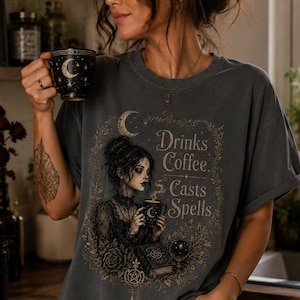 Può includere: Una t-shirt grigio antracite con un'illustrazione grafica di una persona che tiene una tazza di caffè, circondata da rose, un libro e una sfera di cristallo. Il testo recita Drinks Coffee. Casts Spells. Include motivi lunari celestiali.