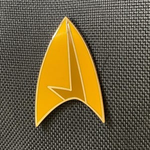 May include: A yellow and gold Starfleet insignia pin. The pin is shaped like a delta with a smaller delta inside. The pin is a popular collectible for fans of the Star Trek franchise.