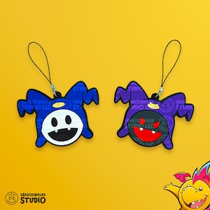 May include: Two Jack Frost character charms, one blue and one purple, with black lanyards. The blue charm has a white face with a black mouth. The purple charm has a black face with a red mouth. The charms are from Dragooneielad Studio.