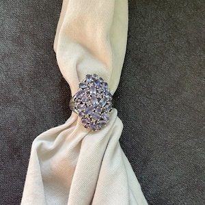 May include: A silver-toned ring featuring a large, oval-shaped cluster of numerous small, light purple, marquise-cut stones. The ring is threaded onto a light beige, ribbed fabric scarf, showcasing the intricate arrangement of the faceted gems.