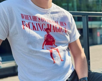 May include: A white t-shirt with a red graphic print that says "When he's on the ball he's f*cking magic". The graphic features a cartoon football player in a red jersey. The person wearing the shirt is sitting on a bench with a black railing in the background.