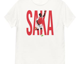 May include: White t-shirt with the name "SAKA" in large red letters. A graphic of a soccer player in a red and white kit is positioned within the letters, celebrating with an arm raised.