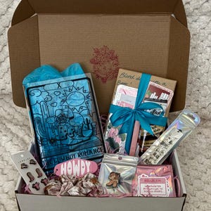 May include: A white box filled with various items, including a book titled "The Cowboy Romance", a pink hair scrunchie, a pink and white "Howdy" sign, a pink and white sticker that says "Hello, I'm obsessed with books", and a clear bag with a pink and white boot pin.