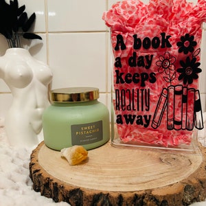 May include: A clear glass jar with a pink and white patterned tissue inside. The jar has a black vinyl decal that reads "A book a day keeps reality away" with a flower and stack of books graphic. A green candle with a gold lid and the label "Sweet Pistachio Scented Candle" sits on a wooden slice.