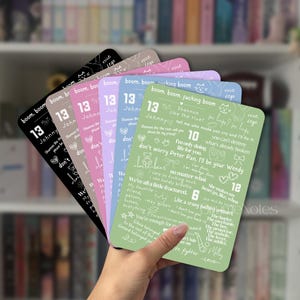 May include: A hand holds a stack of rectangular cards in various pastel colors. Each card features white text, including phrases and numbers like "13" and "Johnny." The cards have a rounded top edge and a matte finish, with a blurred bookshelf in the background.