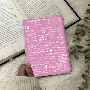 Puede incluir: Una funda de teléfono rosa con texto y garabatos blancos. La funda presenta frases como "boom, boom, fucking boom", "Johnny" y "She makes my heart go, like, whoa". La funda se muestra junto a un libro abierto.