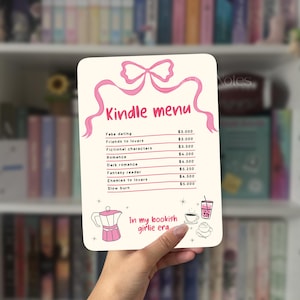 Puede incluir: Un letrero rectangular blanco con un lazo rosa y las palabras "Kindle menu" en rosa. El letrero enumera géneros de libros y precios, incluyendo "Fake dating" y "Romance". La frase "In my bookish girlie era" también está en el letrero.