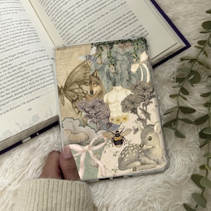 以下が含まれることがあります： 透明な携帯電話ケース。コラージュイラストのデザイン。蝶、オオカミ、蜂、鹿、花、彫刻された胴体が含まれています。「Elegant Jasmine」の文字が見えます。ケースは開いた本の上に置かれています。