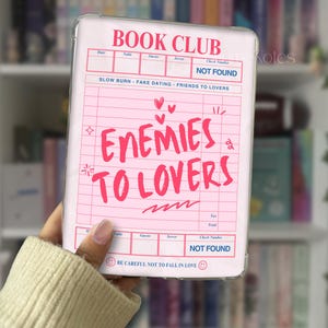 May include: A clear phone case with a pink insert featuring the words "BOOK CLUB" and "ENEMIES TO LOVERS" in red. The design resembles a receipt with lines and boxes. The text "SLOW BURN - FAKE DATING - FRIENDS TO LOVERS" is also visible.