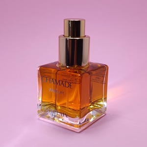 Könnte beinhalten: Ein quadratischer Parfümflakon mit goldenem Verschluss und klarer Glasbasis. Die Flasche enthält eine goldbraune Flüssigkeit und trägt den Aufdruck "CHAMADE". Der Markenname "GUERLAIN PARIS" ist ebenfalls sichtbar.