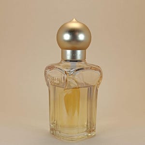 Könnte beinhalten: Eine transparente Parfümflasche aus Glas mit einem goldfarbenen Verschluss. Die Flasche hat eine einzigartige Form mit einem abgerundeten Oberteil und einem rechteckigen Boden. Das Parfüm im Inneren hat eine hellbernsteinfarbene Farbe. Der Markenname "Chopard" ist sichtbar.