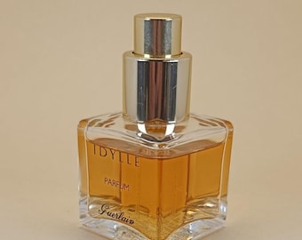 Könnte beinhalten: Ein quadratischer Parfümflakon mit einer goldfarbenen Flüssigkeit. Er hat einen silbernen Verschluss und die Worte "IDYLLE PARFUM Guerlain" sind auf der Vorderseite aufgedruckt. Der Flakon steht vor einem neutralen Hintergrund.